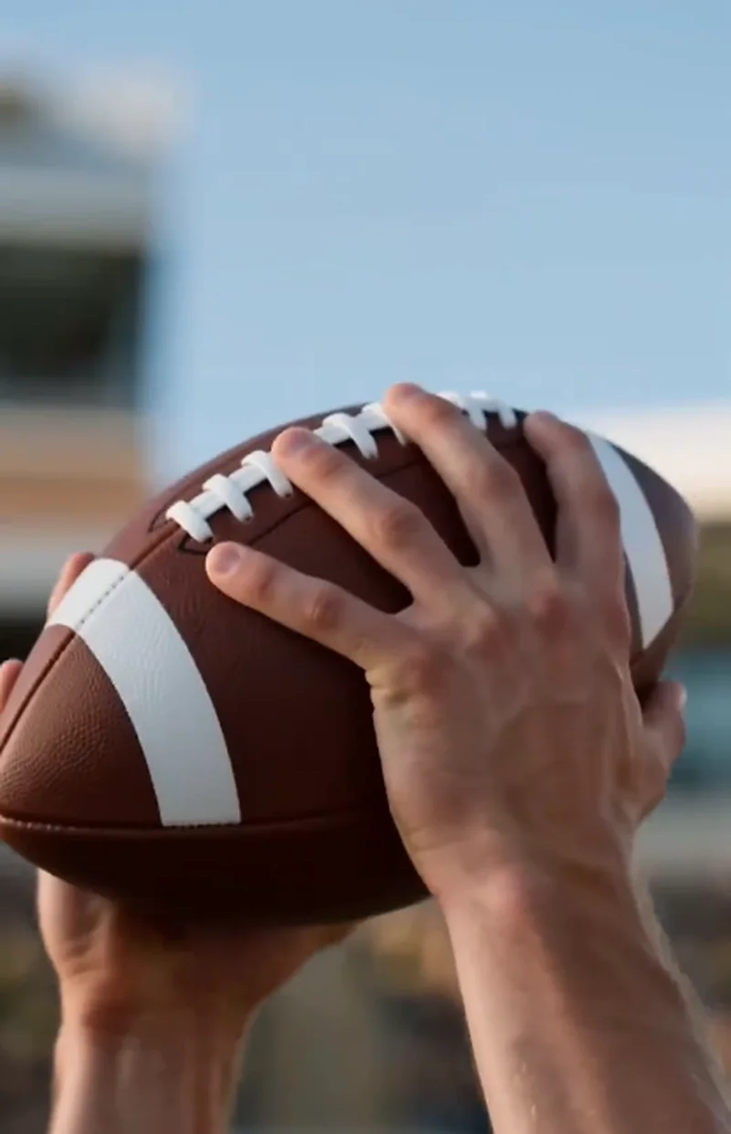 Close-up of hands gripping a brown and white football against a blurred blue sky, ready to pass.