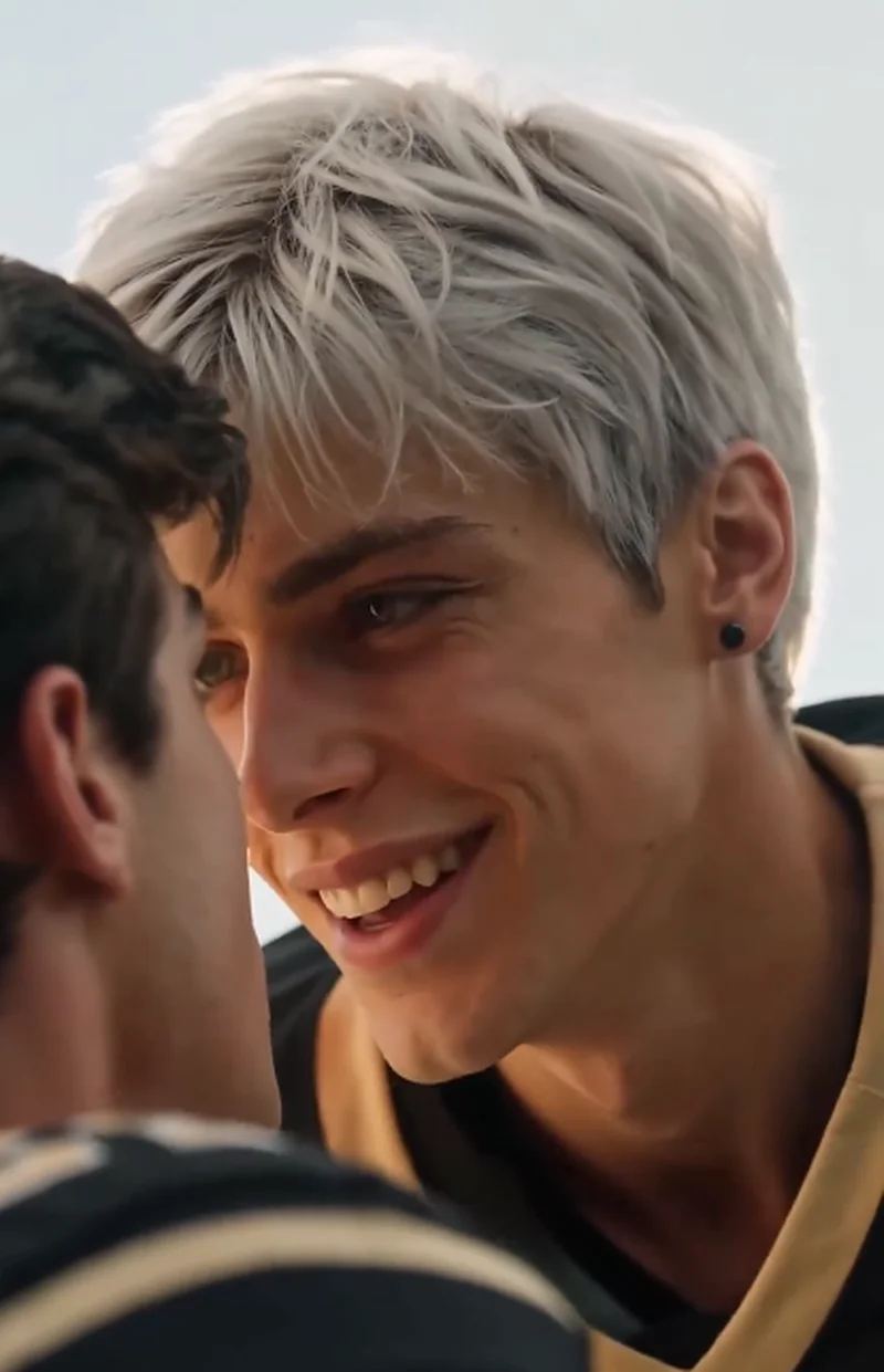 Dylan (silver hair) leans in close to Ethan (dark brown hair) with a playful smile, both in black and gold football uniforms on the field.