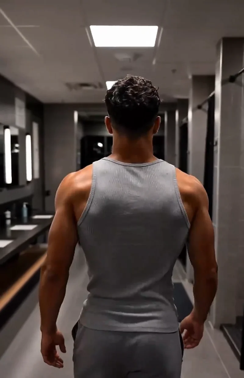Ethan (dark brown hair, gray tank top and sweatpants) walks away down the locker-room aisle, his muscular back and arms in view.