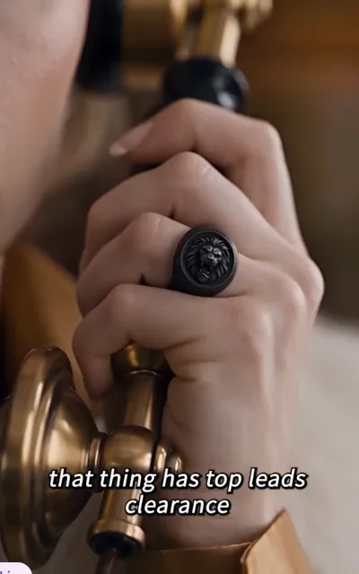 Close-up of a hand holding the brass receiver, black lion-head signet ring on the finger, subtitle: that thing has top leads clearance