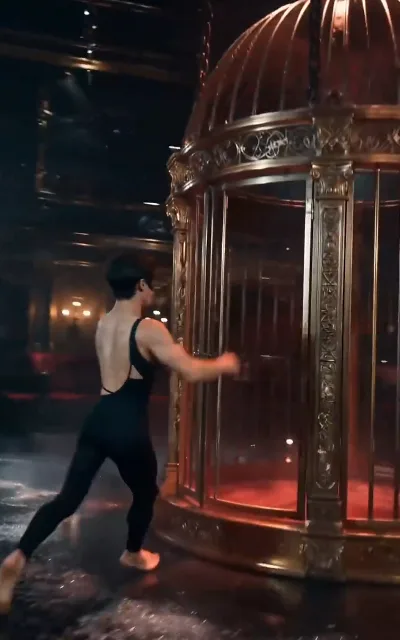 Barefoot dancer pushes an ornate antique brass birdcage glowing red from within, across a flooded stage floor